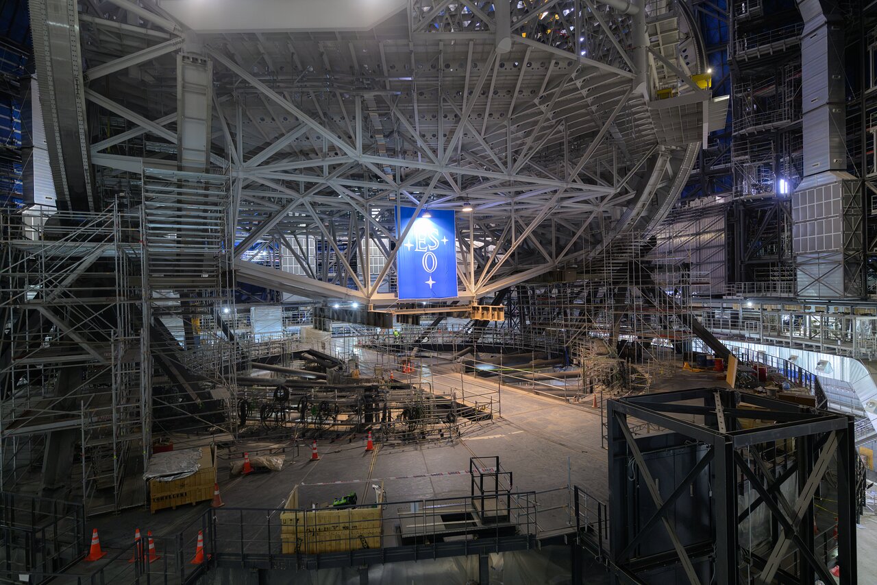 The orange and white cones scattered around the floor of ESO’s Extremely Large Telescope (ELT), currently under construction in the Chilean Atacama Desert, reveal the scale of the future facility. The world’s biggest eye on the sky is progressing fast in preparation for its first light in 2029. A giant ESO flag, put up on the occasion of a visit by the ESO Council in mid June 2026, is seen below the structure.